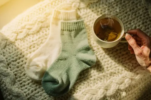 Cream and sage green wool socks on knitted blanket with steaming herbal tea cup in warm morning sunlight